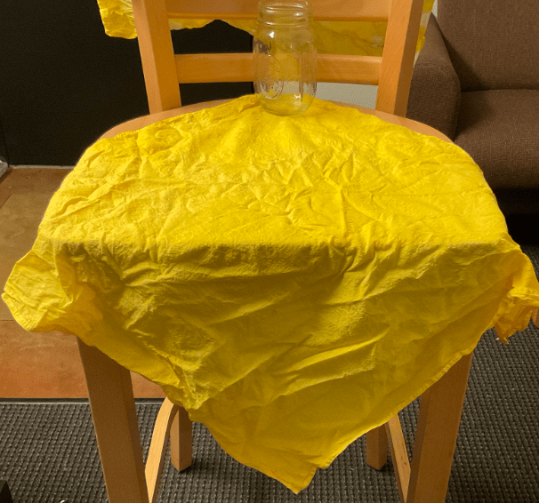 Bright yellow bandana over a chair, being held down by a glass chair; the bandana was dyed with marigold flower